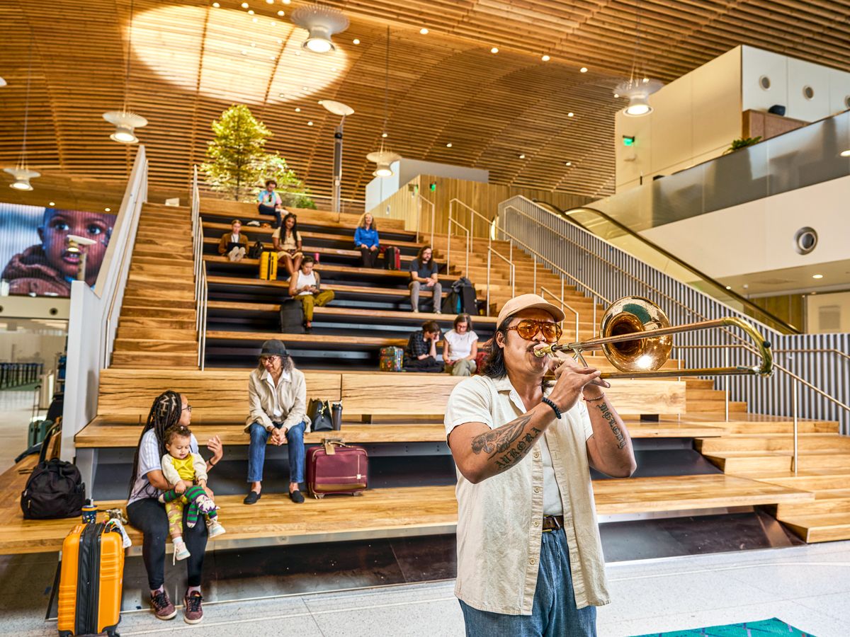 Person playing trumpet next to stairs where passengers sit in Portland International Airport in Oregon