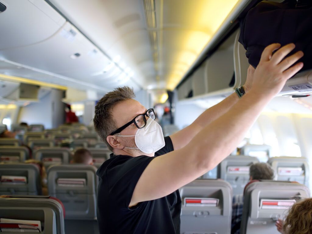 Airline passenger wearing mask stowing bag in overhead bin